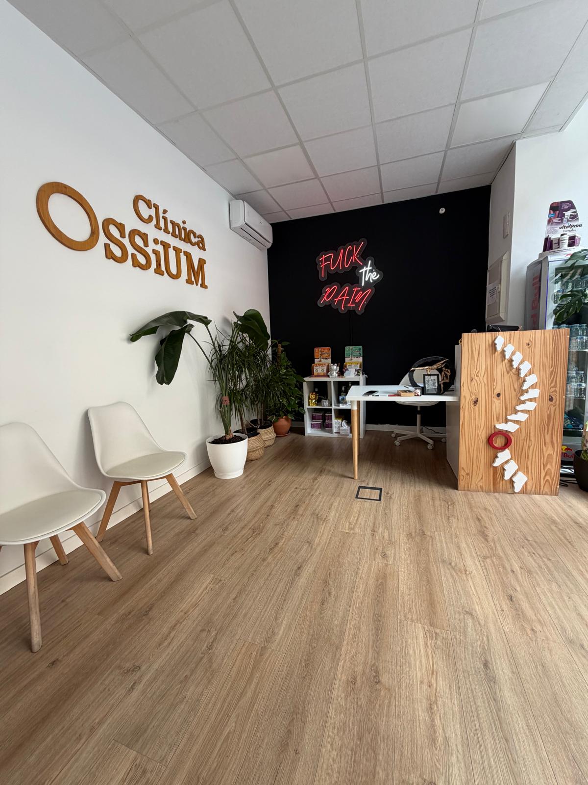 Sala de clínica con sillas blancas, plantas, letrero “Clínica OssiUM” y recepción con neón “Fuck the PATIN”.